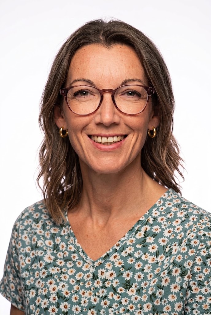 Retrato de una mujer sonriente con gafas y pendientes, de pelo castaño ondulado hasta los hombros, vestida con una camisa de flores verdes sobre fondo blanco.